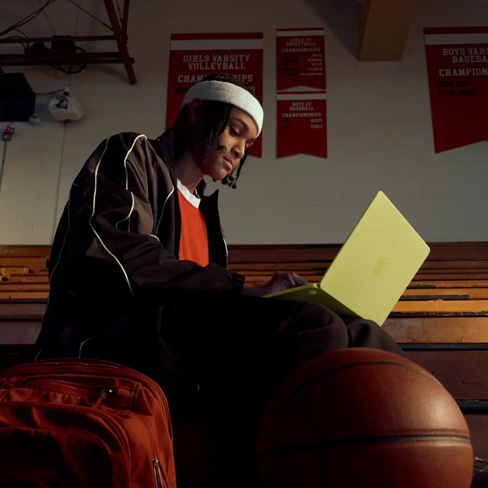 Person sitting on a bench with a laptop, surrounded by sports equipment in a gym.